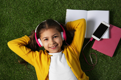 Photo of Cute little girl listening to audiobook on grass, top view