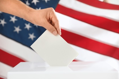 Photo of Man putting ballot paper into box and American flag on background, closeup