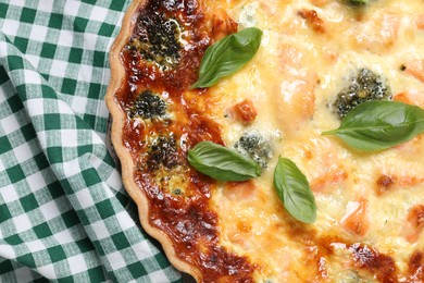 Photo of Delicious homemade quiche with salmon, broccoli and basil leaves on green checkered cloth, top view
