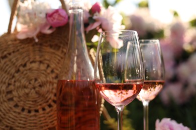 Photo of Bottle and glasses of rose wine near straw bag with beautiful peonies in garden, closeup