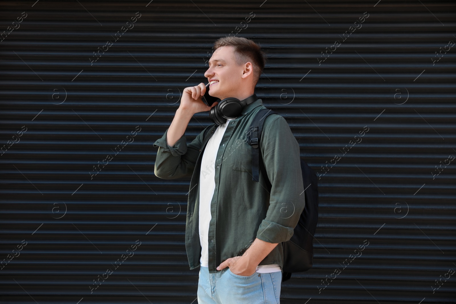 Photo of Smiling man with headphones talking on smartphone near shutters outdoors