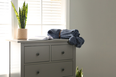 Grey chest of drawers with houseplant, book and blanket near window in room