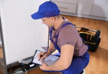 Photo of Male technician with clipboard examining refrigerator in kitchen