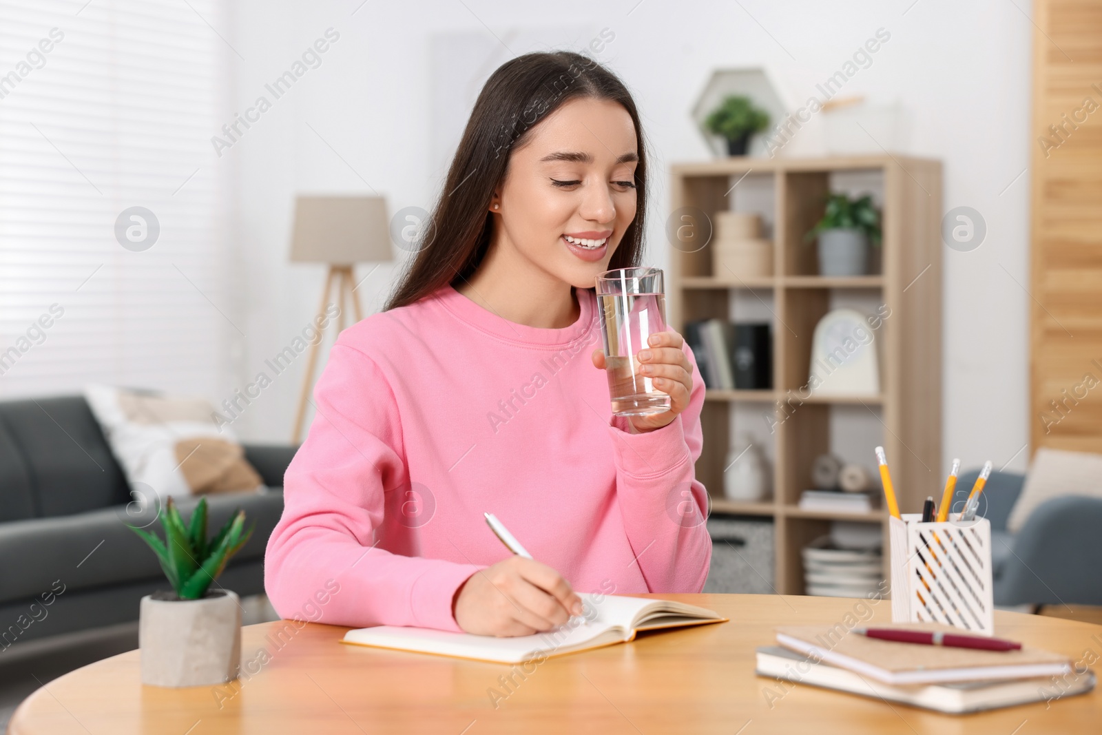 Photo of Young woman with glass of water writing in notebook at wooden table indoors