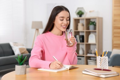 Young woman with glass of water writing in notebook at wooden table indoors