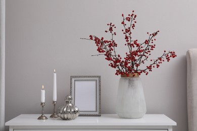 Photo of Hawthorn branches with red berries in vase, candles and frame on table indoors