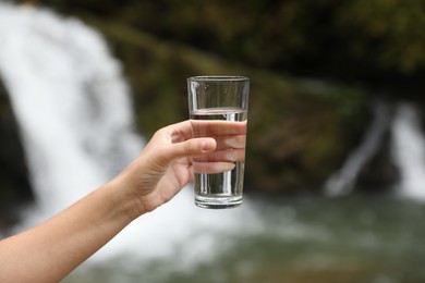 Photo of Woman holding glass of fresh water near waterfall outdoors, closeup