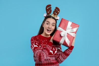 Photo of Surprised young woman in Christmas sweater and reindeer headband holding gift on light blue background