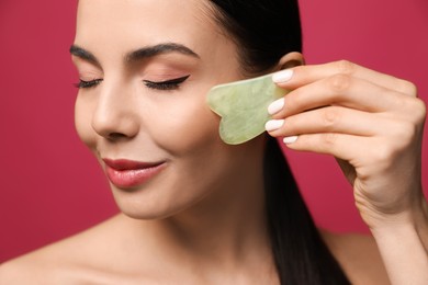 Photo of Beautiful young woman doing facial massage with gua sha tool on pink background, closeup