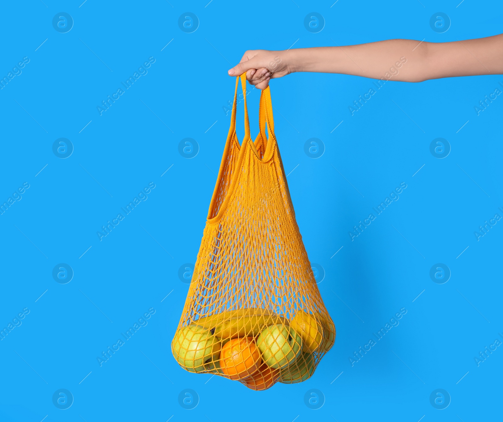 Photo of Woman holding net bag with fruits on light blue background, closeup