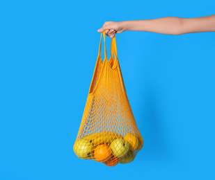 Woman holding net bag with fruits on light blue background, closeup