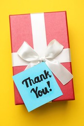 Photo of Gift box and light blue paper note with phrase Thank You on yellow background, top view