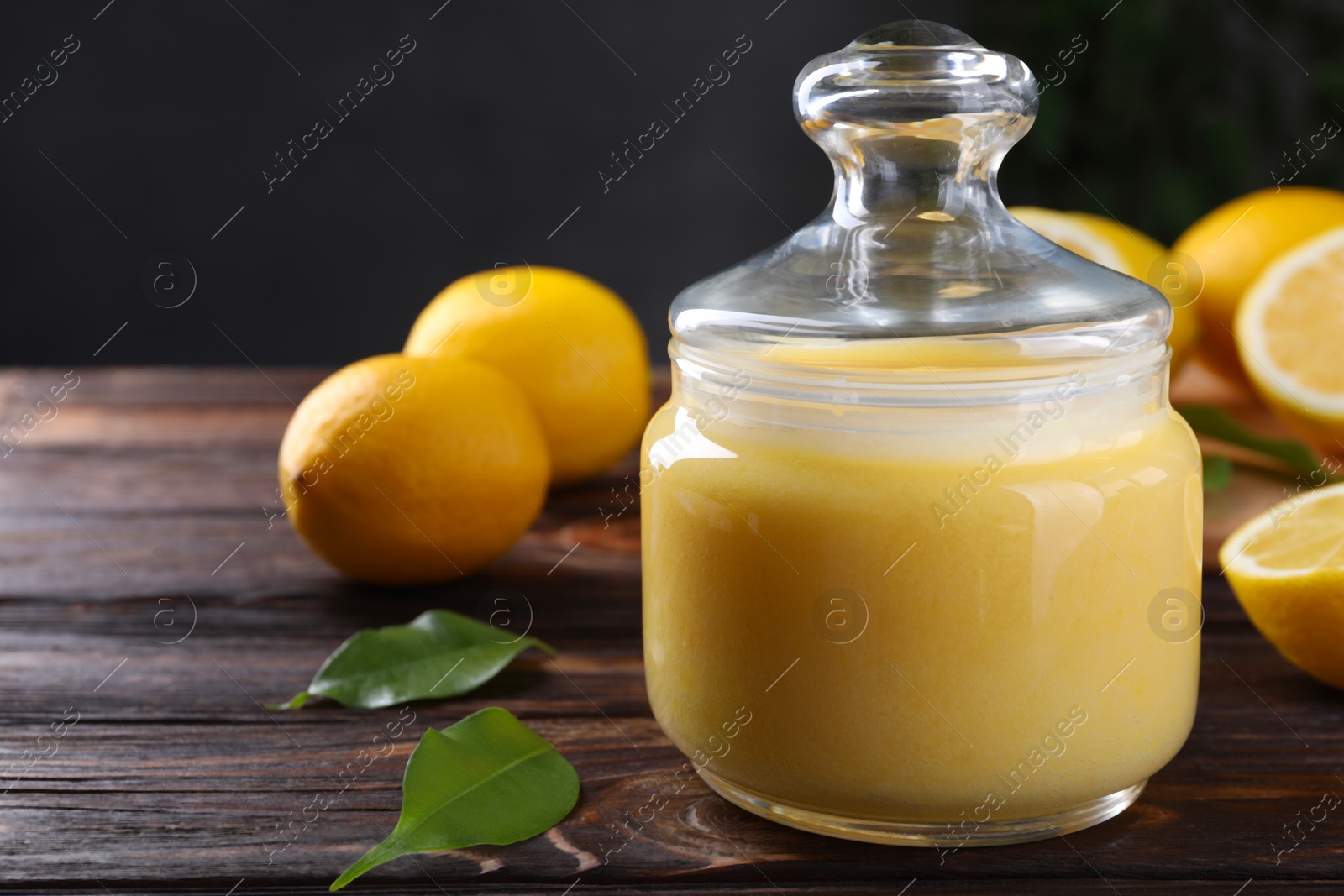 Photo of Delicious lemon curd in glass jar, fresh citrus fruits and green leaves on wooden table