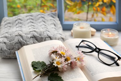 Open book with chamomile flowers as bookmark, scented candles and glasses on white wooden table near window, closeup. Space for text