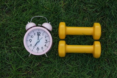 Photo of Alarm clock and dumbbells on green grass, flat lay. Morning exercise
