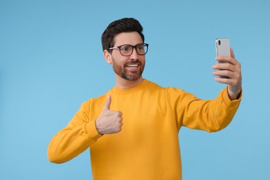Photo of Smiling man taking selfie with smartphone and showing thumbs up on light blue background