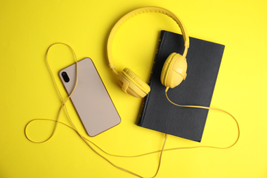 Book, modern headphones and smartphone on yellow background, flat lay