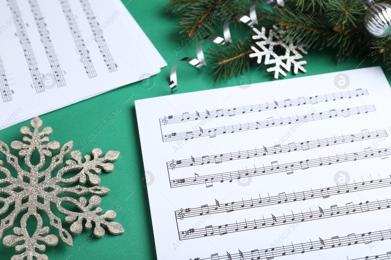 Photo of Christmas music sheets near fir tree branches and decorative snowflakes on green background, closeup