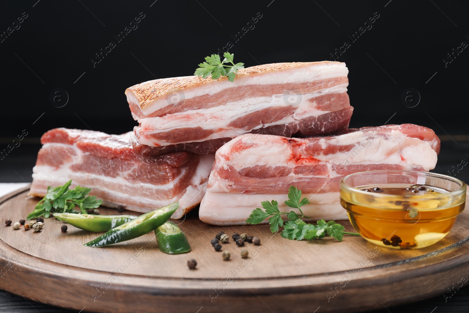 Photo of Pieces of raw pork belly, chili pepper, peppercorns, oil and parsley on black wooden table, closeup