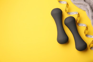 Dumbbells, measuring tape and towel on yellow background, flat lay. Space for text