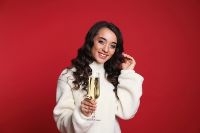 Beautiful young woman in warm sweater holding glass of champagne on red background. Christmas party