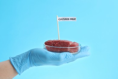 Photo of Scientist holding Petri dish with lab grown beef labeled Cultured Meat on light blue background, closeup