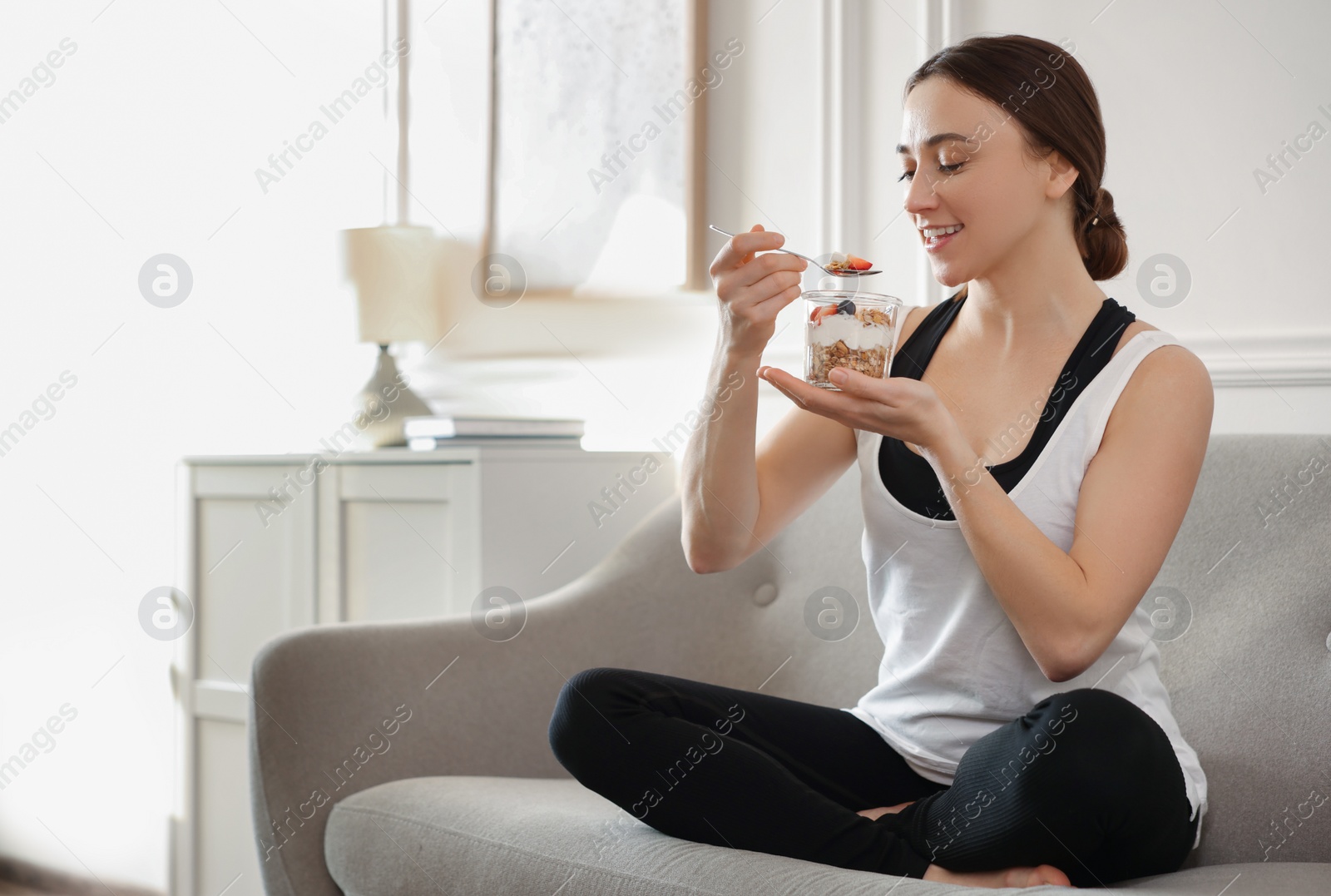 Photo of Woman eating tasty granola with fresh berries and yogurt on sofa at home