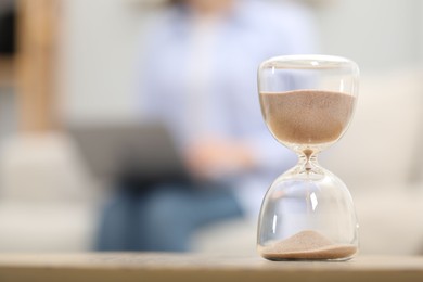 Photo of Hourglass with flowing sand on desk. Woman using laptop indoors, selective focus