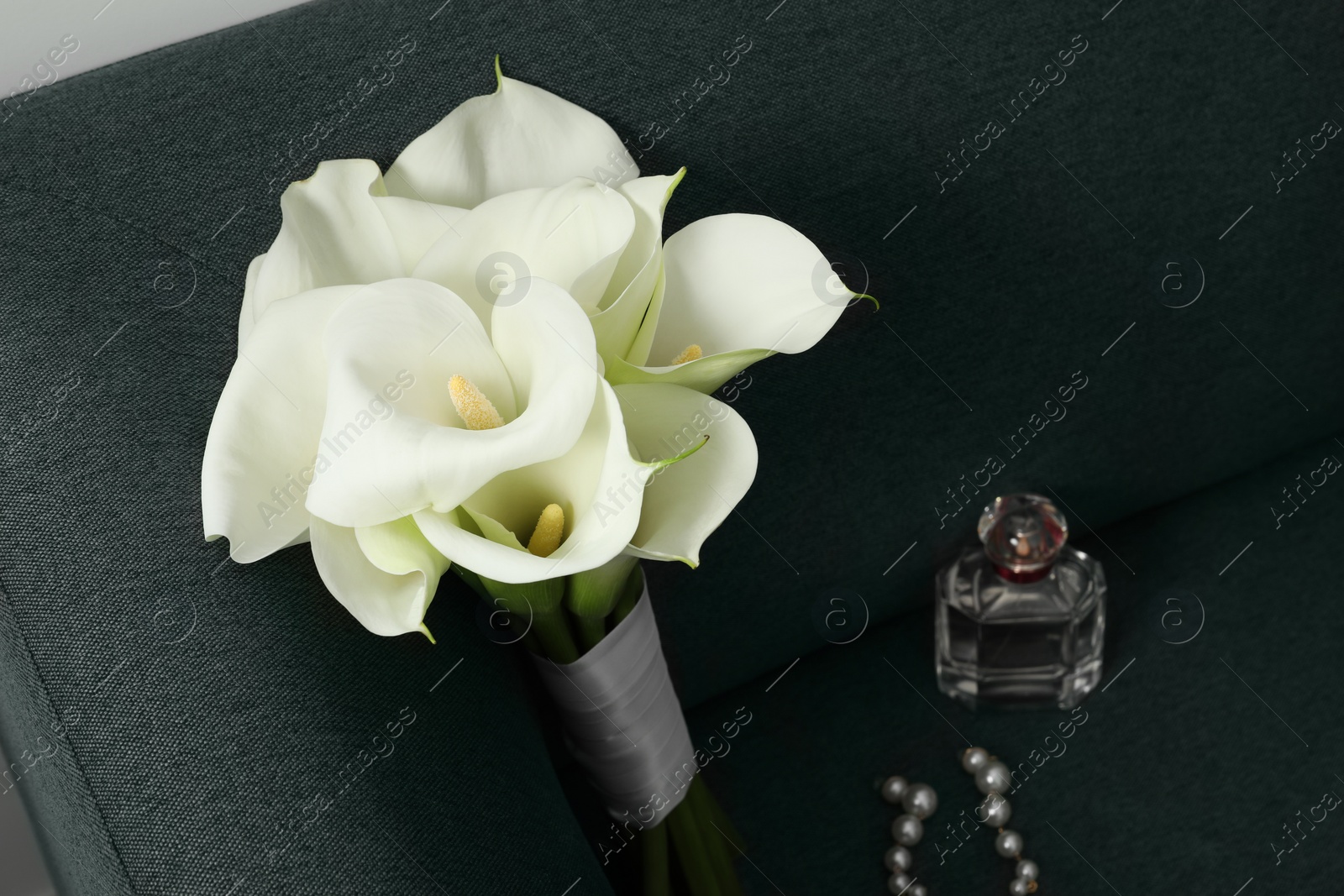 Photo of Beautiful calla lily flowers tied with ribbon, bottle of perfume and earrings on sofa