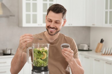 Photo of Happy man adding chia seeds into blender with ingredients for smoothie in kitchen