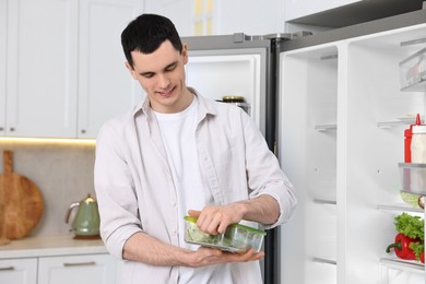 Happy man holding container with vegetables near refrigerator in kitchen