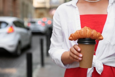 Photo of Woman holding tasty croissant and cup of coffee on city street, closeup. Space for text