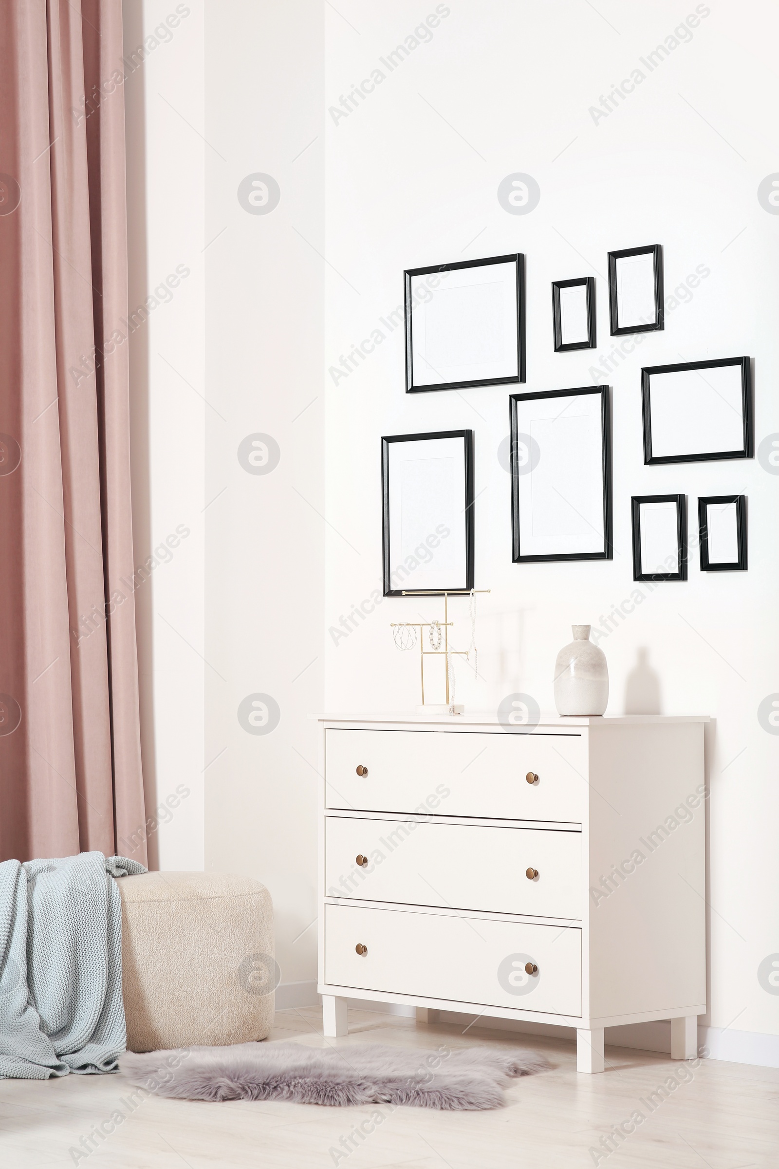 Photo of Empty frames hanging on white wall, chest of drawers and ottoman indoors