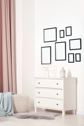 Photo of Empty frames hanging on white wall, chest of drawers and ottoman indoors