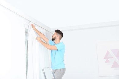 Photo of Young handsome man hanging window curtain in room