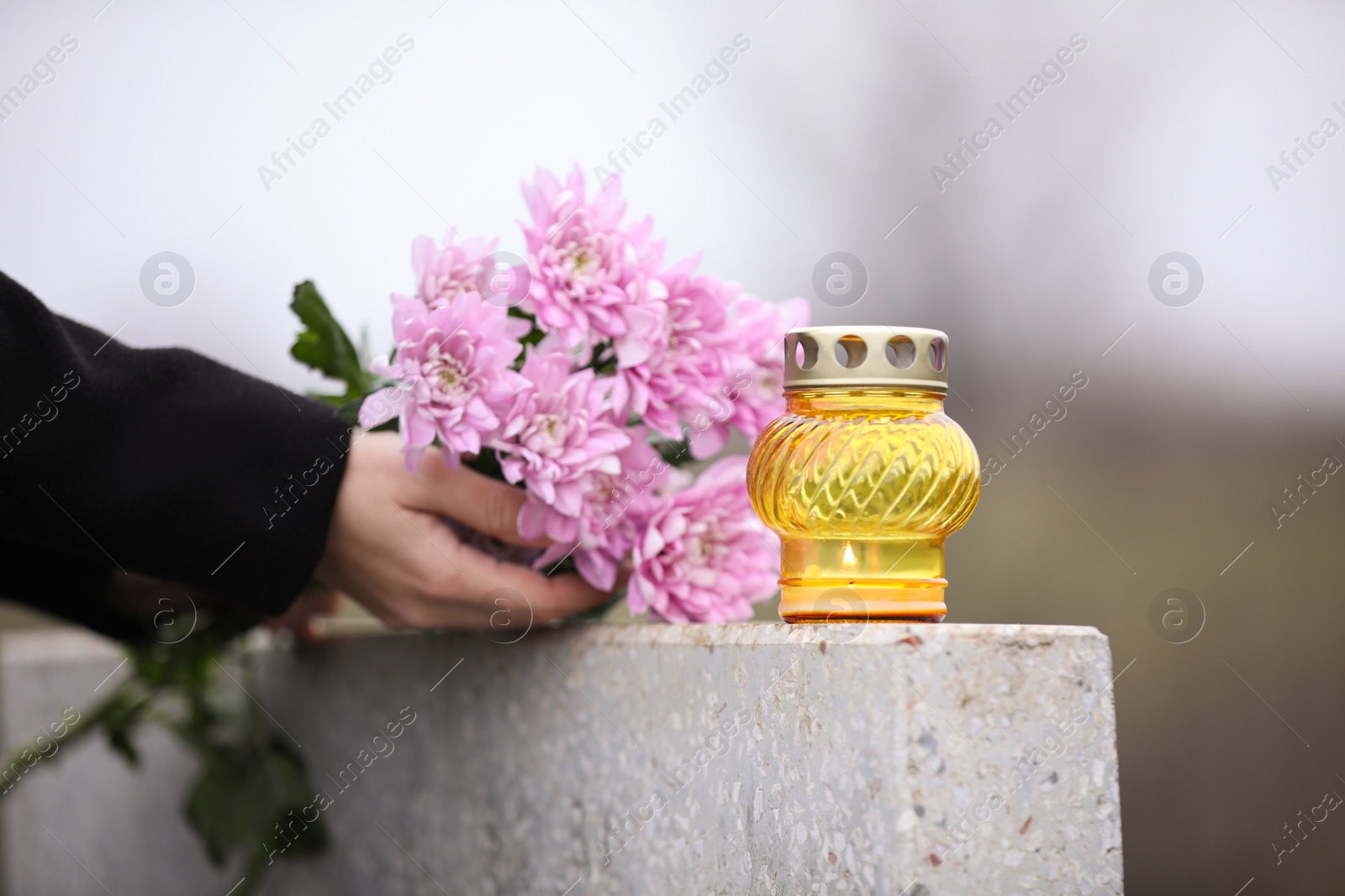 Photo of Woman holding chrysanthemum flowers near grey granite tombstone with candle outdoors, closeup. Funeral ceremony