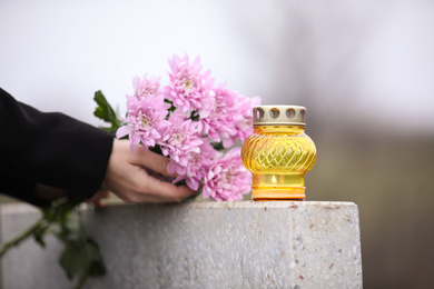 Woman holding chrysanthemum flowers near grey granite tombstone with candle outdoors, closeup. Funeral ceremony