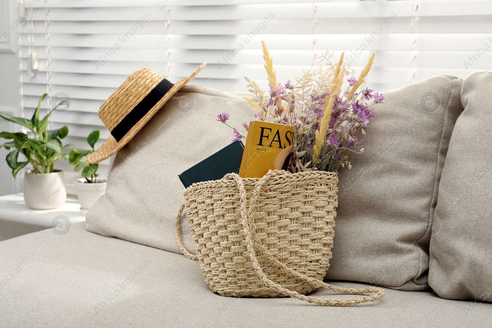 Photo of Stylish beach bag with wildflowers, magazine and book on sofa in room