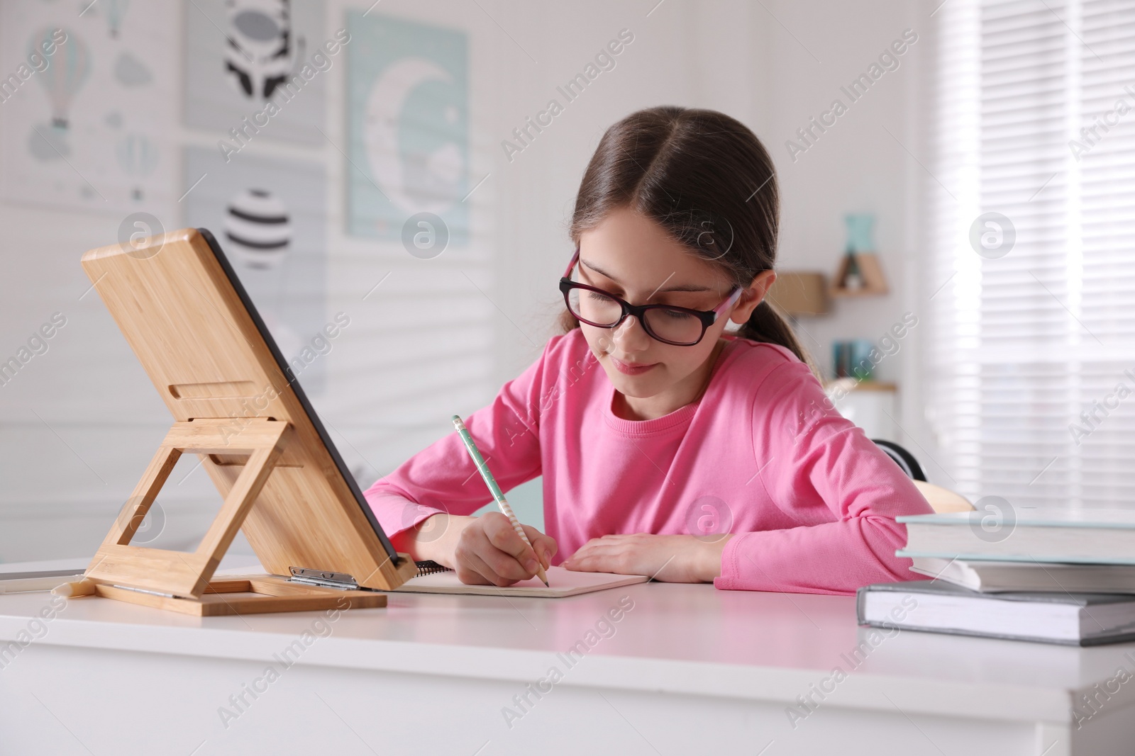 Photo of Little girl doing homework with tablet at table in room