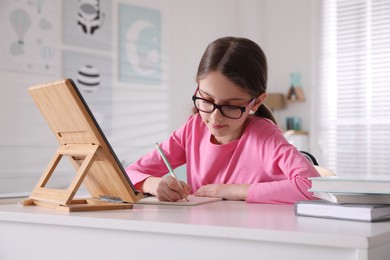 Photo of Little girl doing homework with tablet at table in room