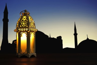 Image of Decorative Arabic lantern on wooden surface and silhouette of mosque at sunset on background, space for text