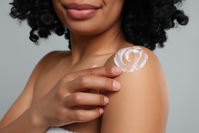 Young woman applying body cream onto shoulder on grey background, closeup