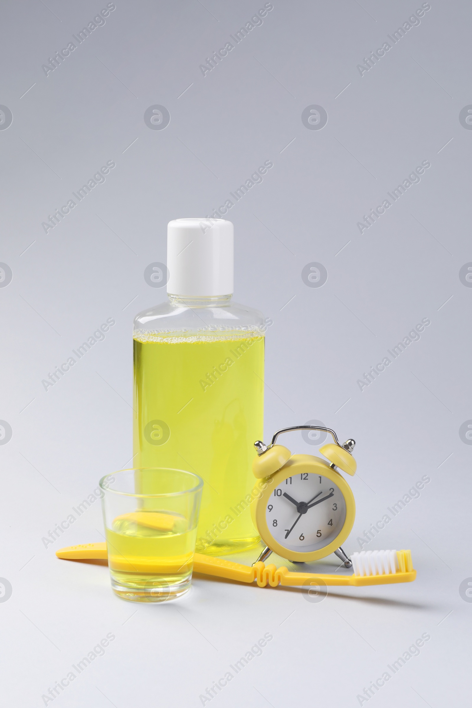 Photo of Fresh mouthwash in bottle, glass, toothbrush and alarm clock on grey background