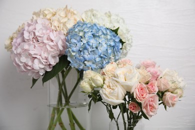Beautiful hydrangea and rose flowers in vases near white wall, closeup