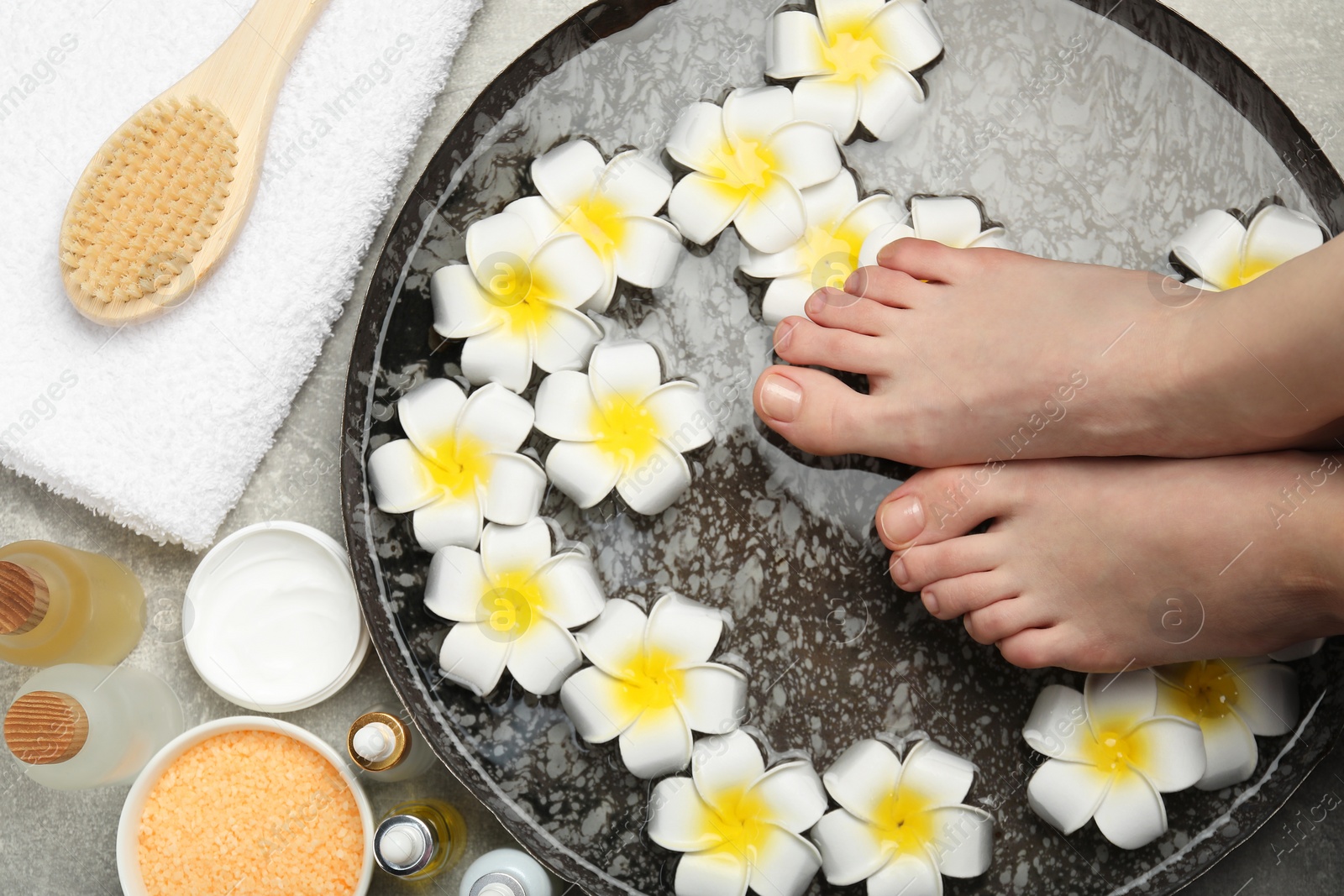 Photo of Woman soaking her feet in bowl with water and flowers on light grey floor, top view. Spa treatment