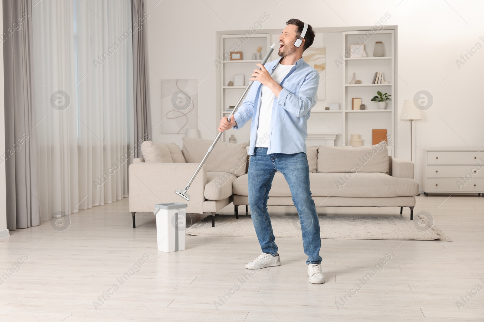 Photo of Enjoying cleaning. Happy man in headphones with mop singing while tidying up at home