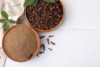 Photo of Aromatic clove powder and dried buds in bowls on white tiled table, flat lay. Space for text