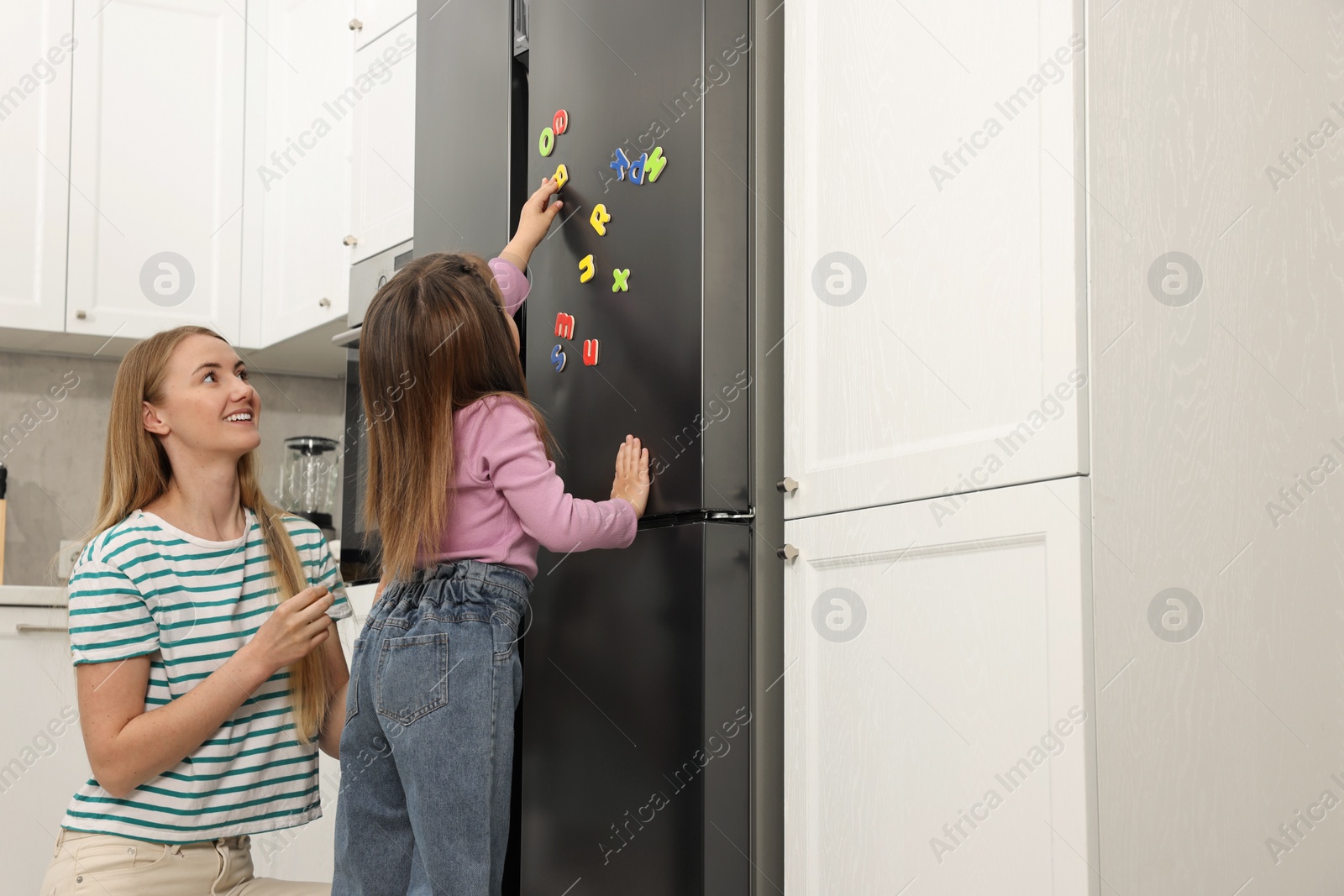 Photo of Mom and daughter putting magnetic letters on fridge at home. Learning alphabet