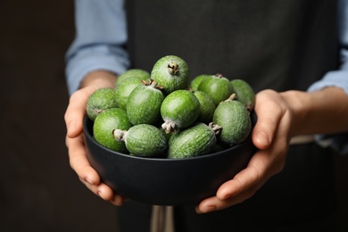 Photo of Woman holding fresh green feijoa fruits in bowl on dark background, closeup