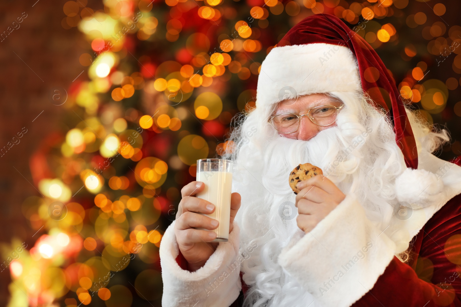 Photo of Santa Claus with milk and cookie near Christmas tree indoors